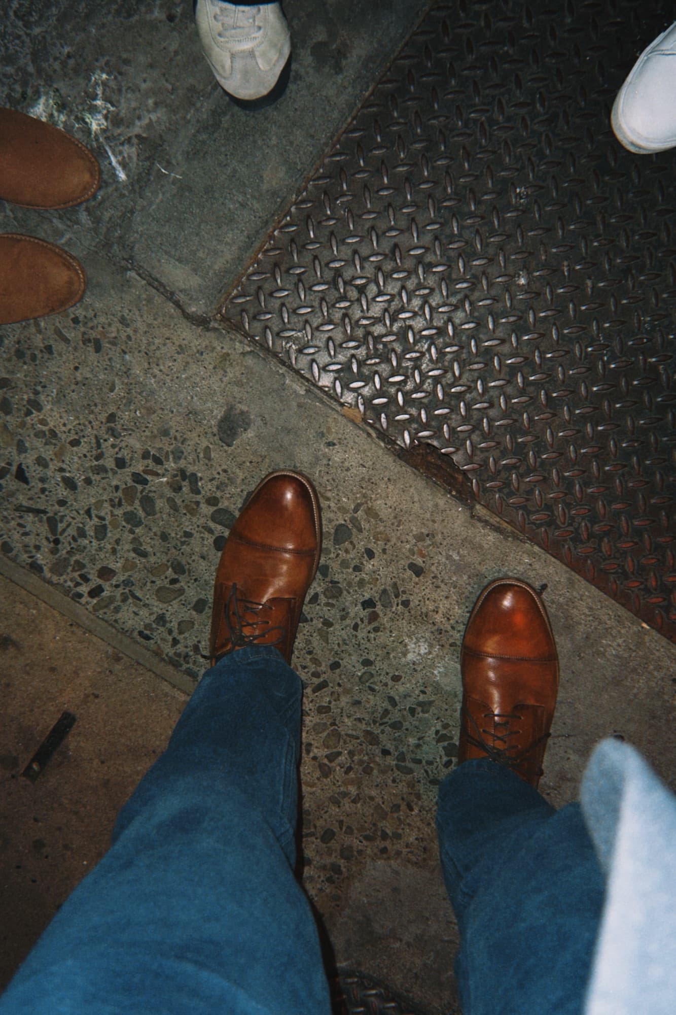Looking down at brown leather boots and jeans, standing near other people's shoes on textured pavement