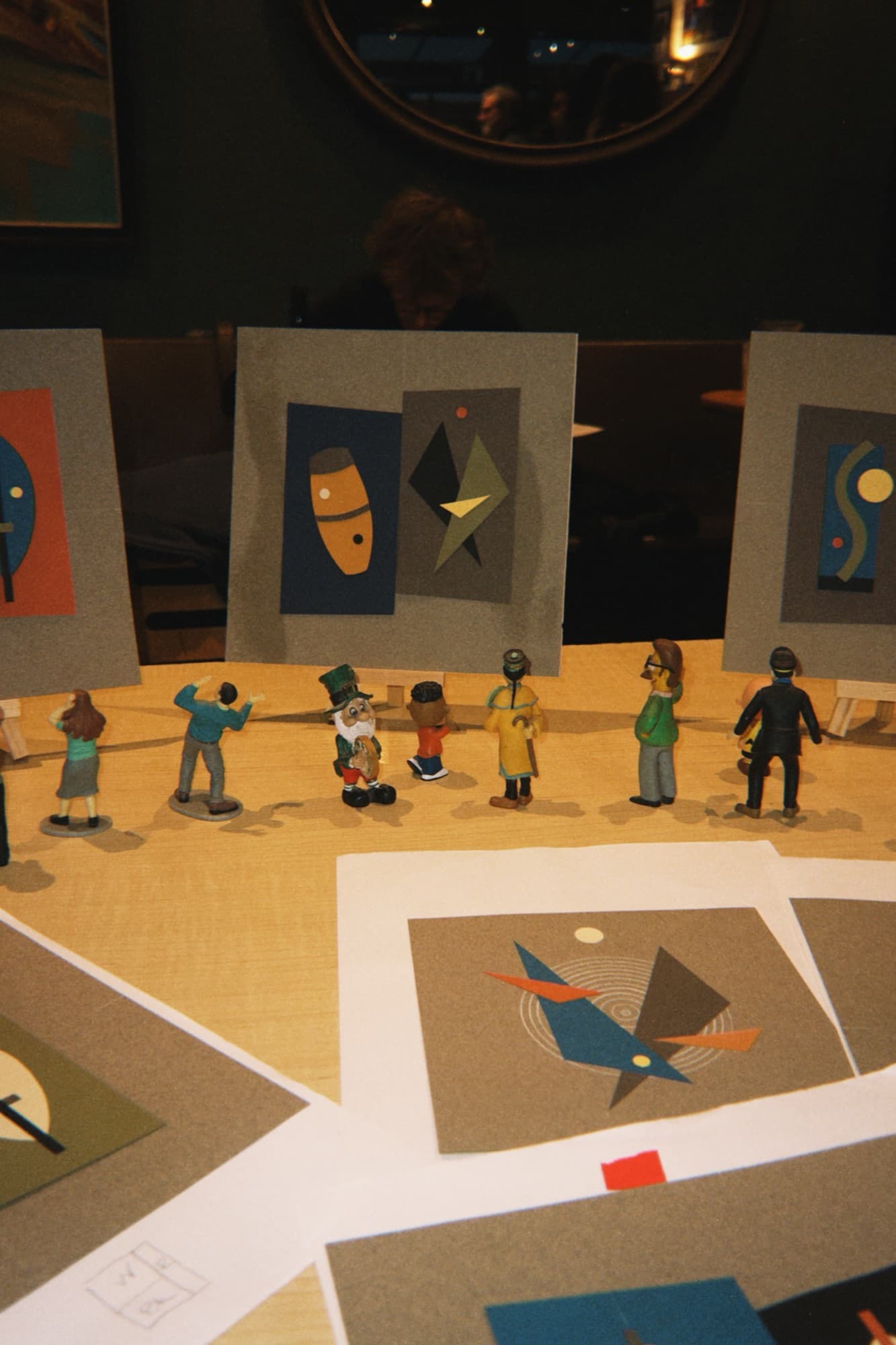 Miniature painted figurines arranged like gallery visitors viewing small abstract art prints
