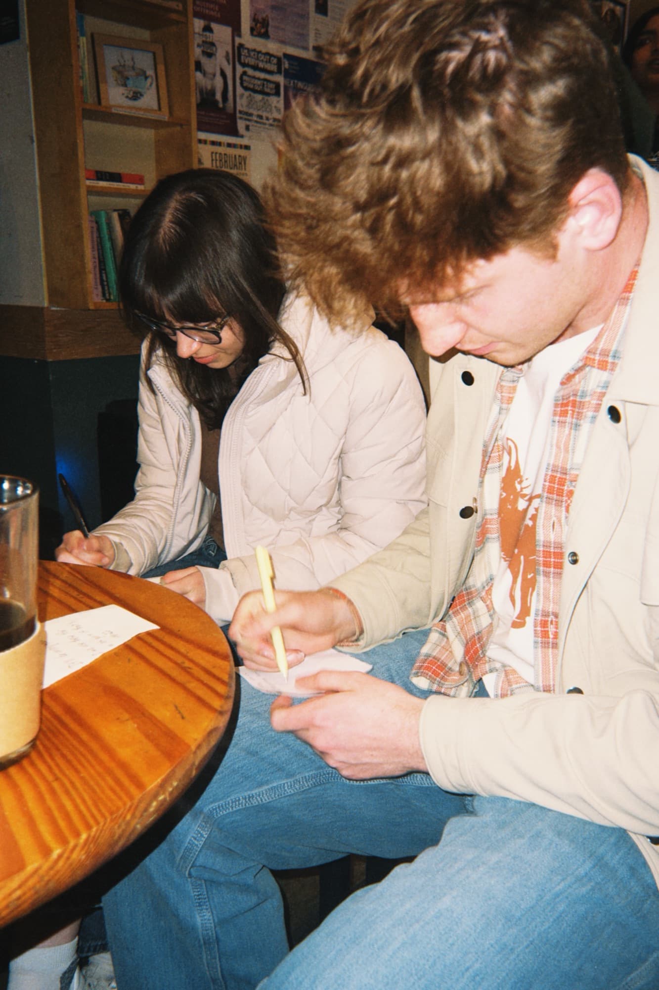 A woman with glasses and a man in a tan jacket writing on paper at a small round cafe table