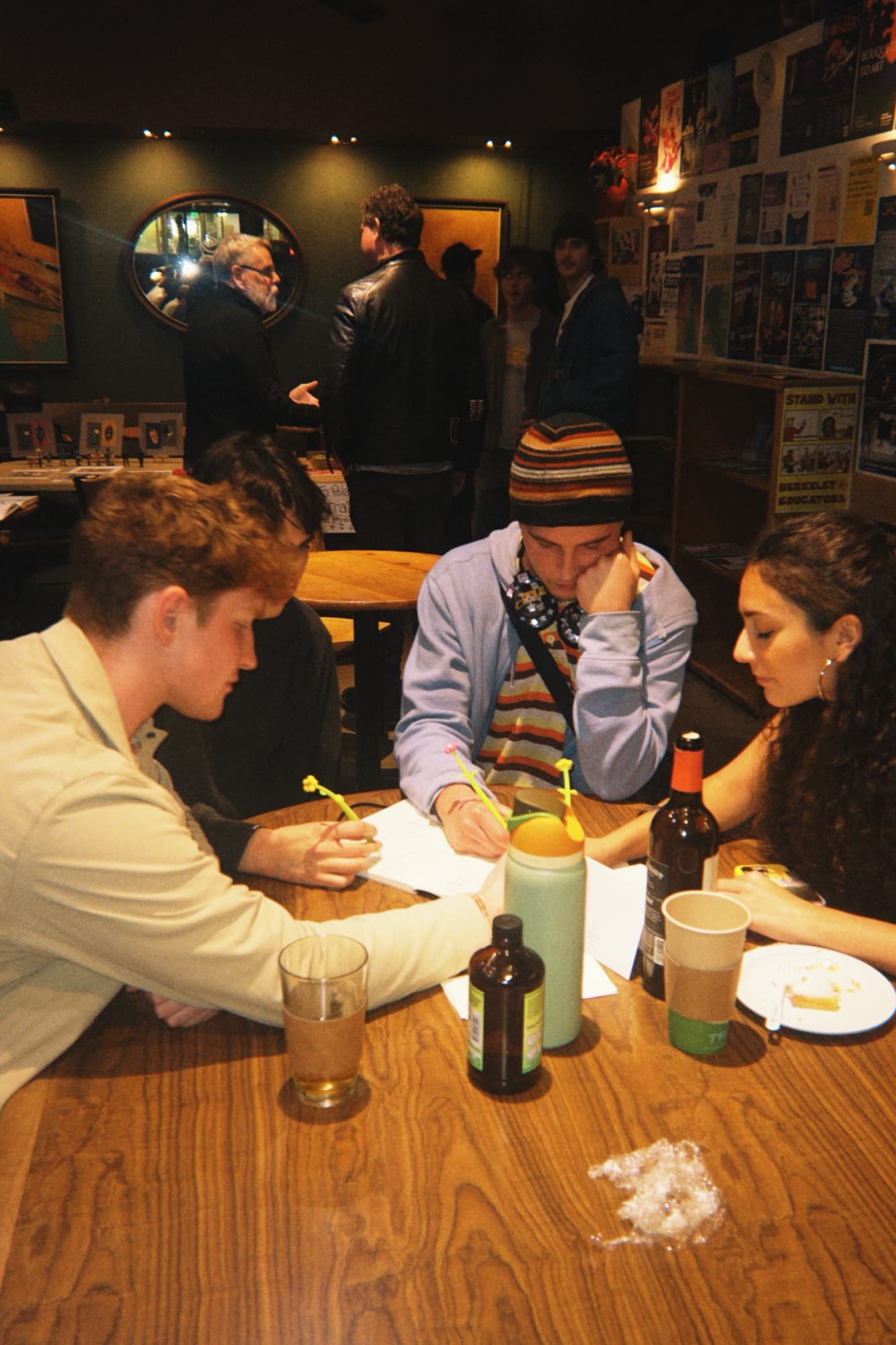 Four people at a wooden bar table writing on paper, with drinks and toy flower pens nearby