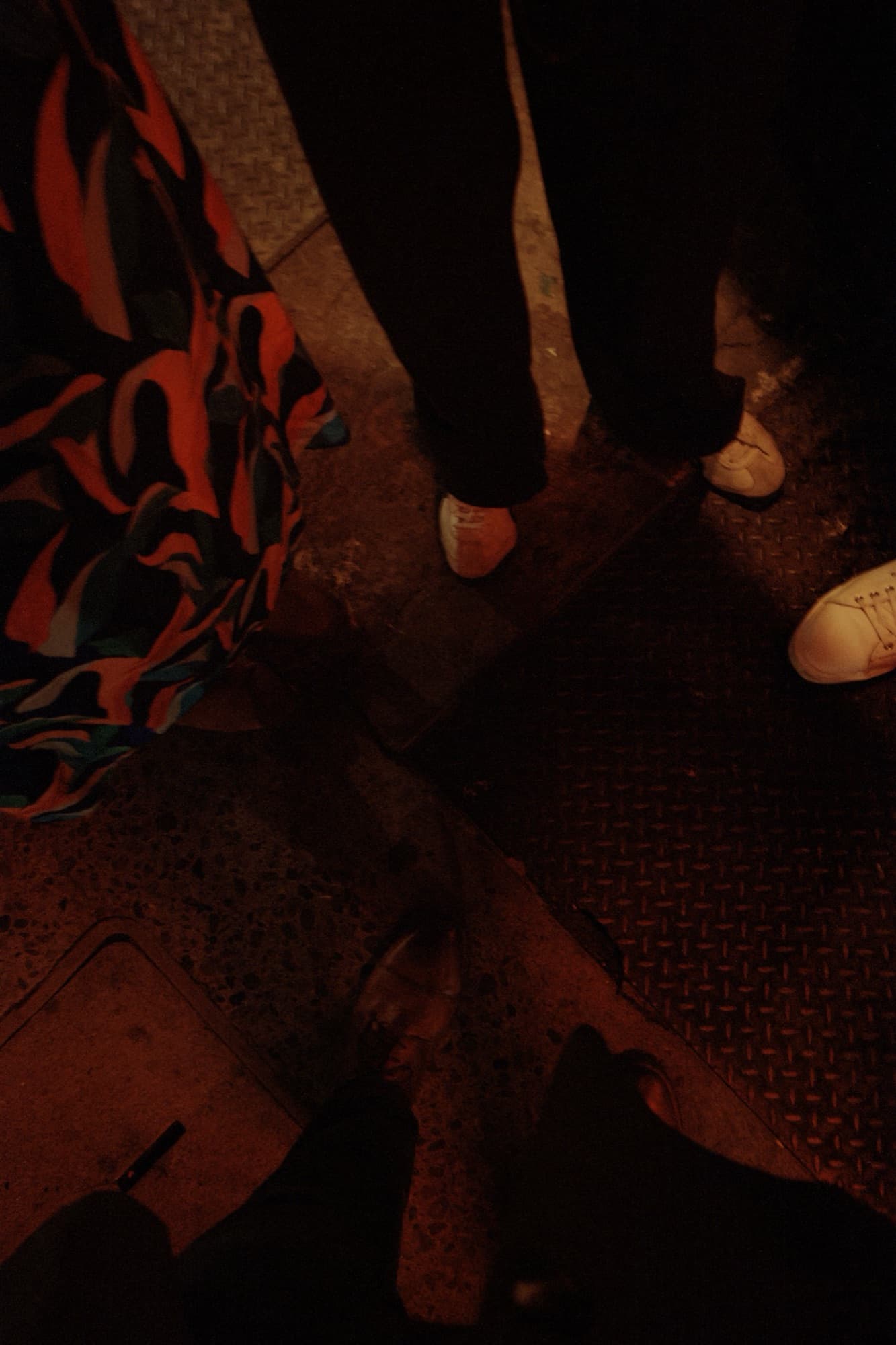 Dark overhead shot of several people's feet and shoes, one wearing a colorful patterned skirt