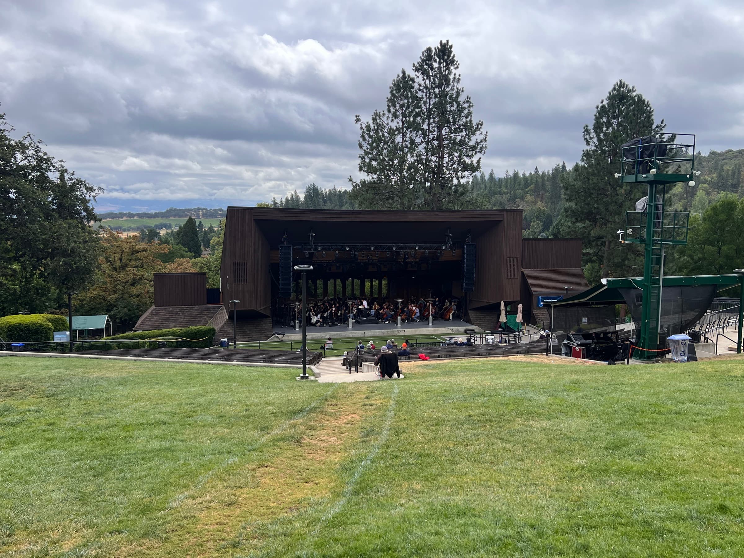 An outdoor amphitheater stage with a seated orchestra, empty bench seating, and cloudy hills beyond