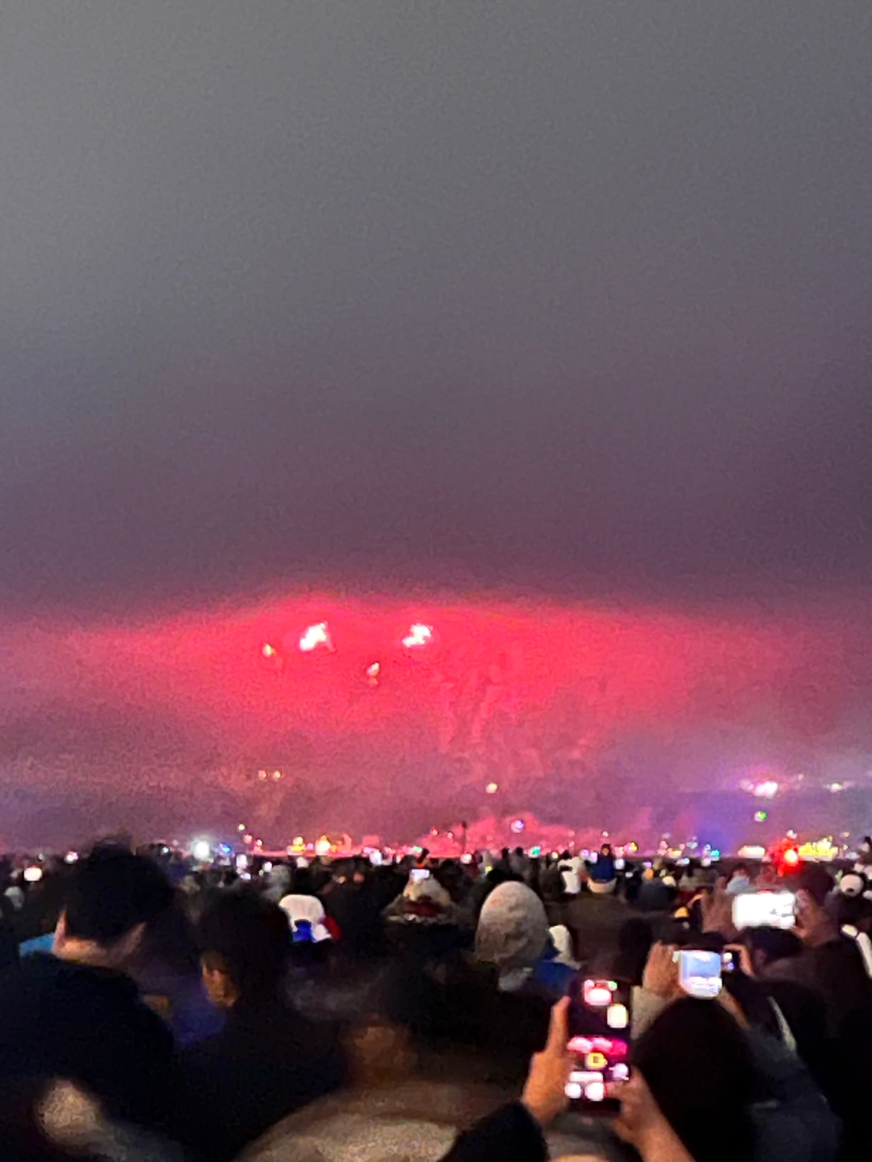 A blurry nighttime crowd holds up phones toward red stage lighting and haze