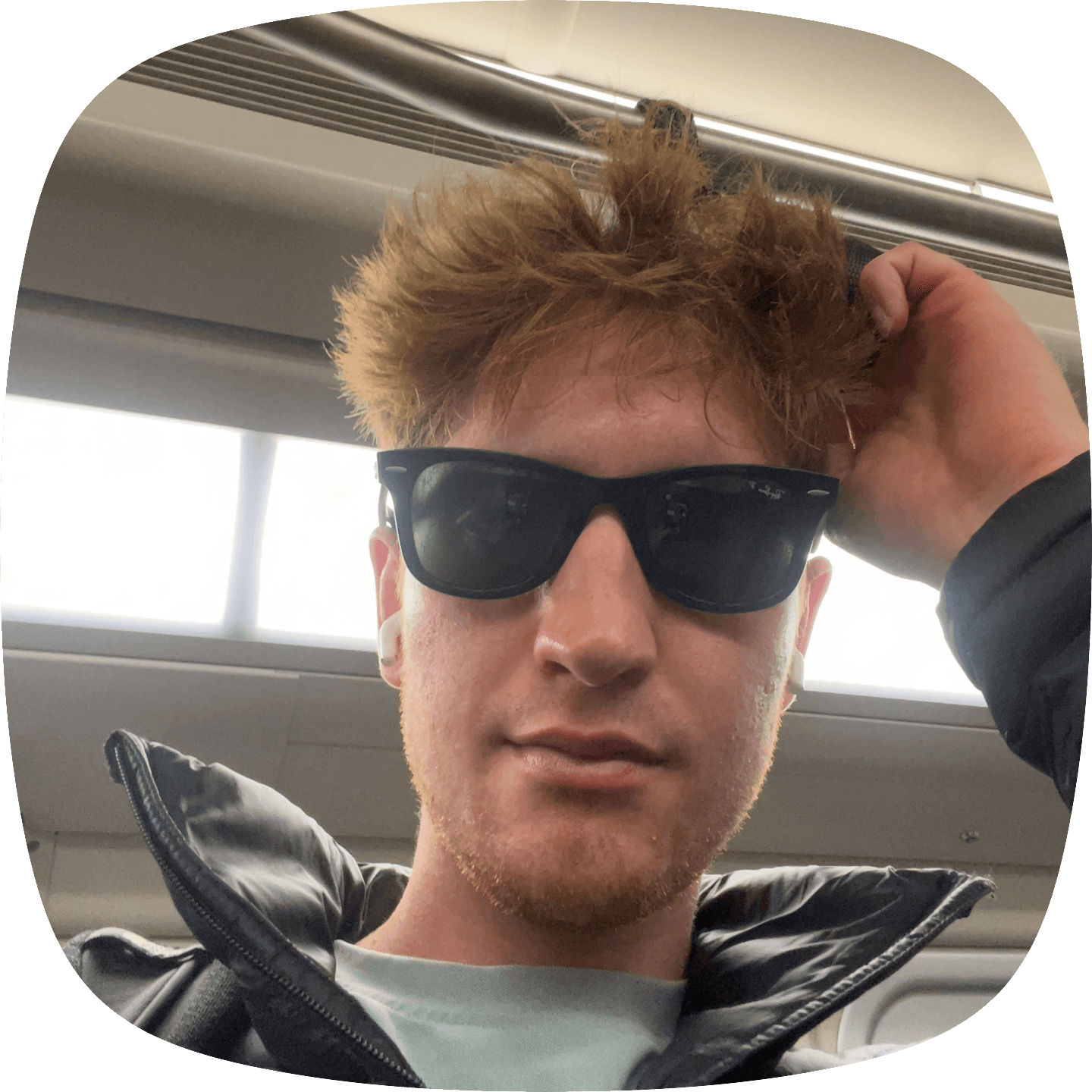 A young man with reddish hair and sunglasses takes a selfie while holding an overhead rail on a train