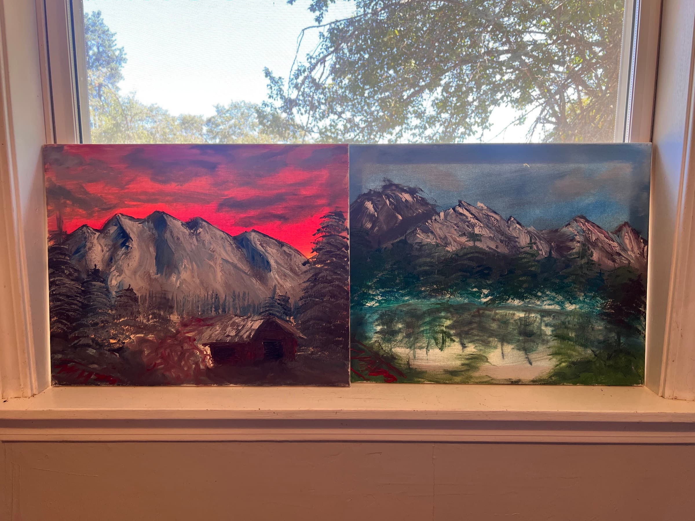 Two landscape paintings on a windowsill, one a red sunset over snowy peaks and a cabin, one a lake