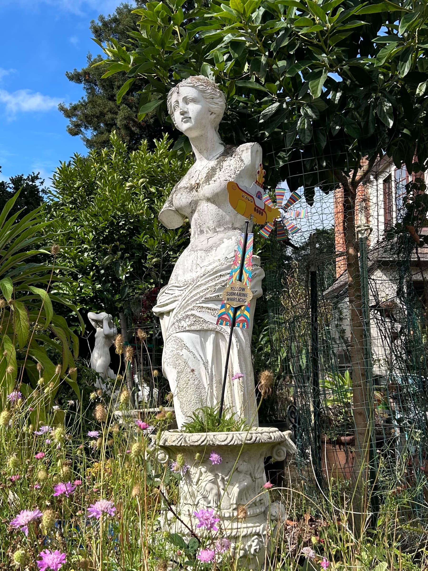 A weathered stone statue of a classical female figure in a garden, decorated with a colorful pinwheel